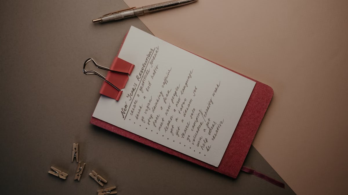 Top view of notebook with handwritten New Year's resolutions, pen, and clips on a desk.